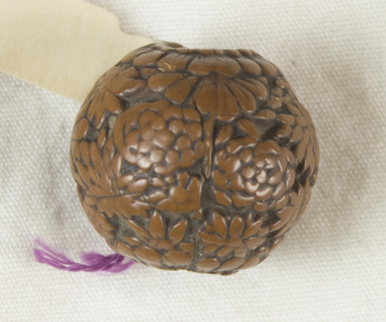 Hearst Museum object titled Bead, accession number 9-12488, described as Japan. Ojime: carved fruit pit or nut. Allover design of floral motif. Cord hole through middle. Diameter 1.6 centimeters.