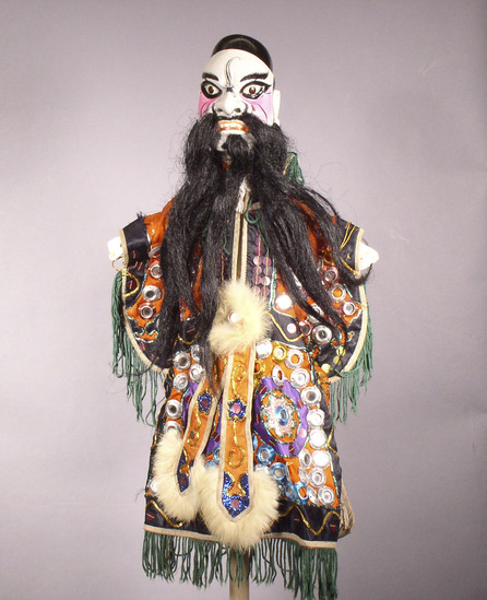 Hearst Museum object titled Hand puppet, accession number 9-22231, described as Puppet, hand.  Clown  A. Head: carved wood, white with pink, orange, green and black features, painted black skull cap, black beard and moustache  B. Body: white cloth.  Boots:  wood, black.  C.  Robe: orange cloth, sequins, metal mirrors, green fringe, white rabbit fur trim  45cm x 30 cm (17 11/16 x 11 13/16 in)