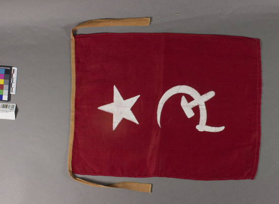 Hearst Museum object titled Flag, accession number 7-6676, described as FLAG:  Red cotton printed in center with five pointed star over hammer and sickle in stiff white paint.  Top bound w. narrow tan band extending 7 inches on each side as ties.  Cotton.  Printing, painting.  Red, white and tan.  2 hemmed edges, 1 selvedge.  16 inches by 12 ⅛ inches.