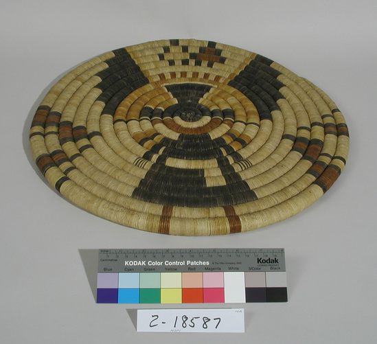 Hearst Museum object titled Plaque, accession number 2-18587, described as Basketry, coiled.  Black, white, brown and yellow.  Full length Kachina figure on plaque; black body, partially black headdress (with added small coil) with triangular elements on each side at head; triangular or terraced elements on each side of black body, white inverted "L" on black body; hands black, feet brown and yellow.