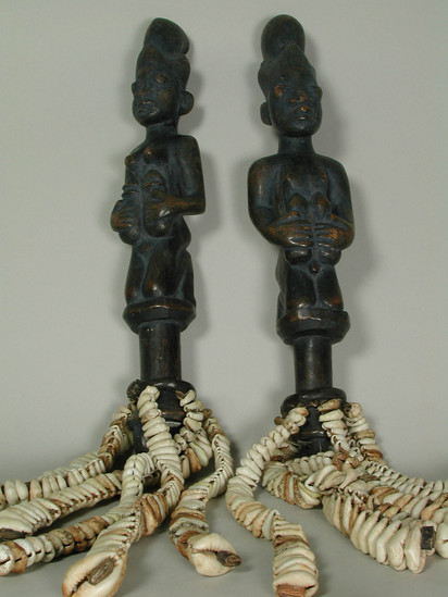 Hearst Museum object titled Figures, accession number 5-15796a,b, described as figures, wood, handcarved; dark polished, each with handle and strands of cowrie shells suspended from leather covered ring near proximal end; both figures kneeling, and wearing headdress; a) male, with hands below knees, b) female, holding flasks in each hand against chest;  L. ea. 34.0 cm. Has bag of shell fragments.