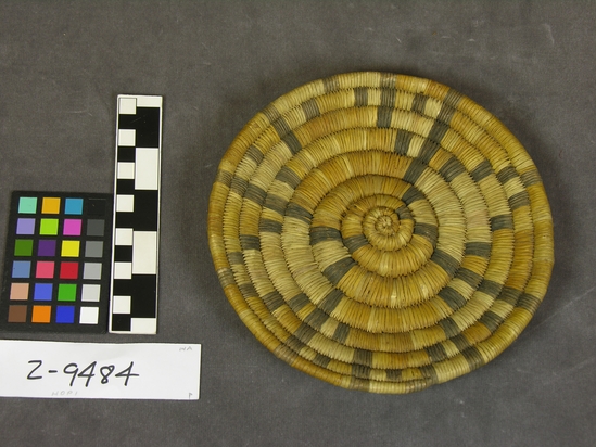 Hearst Museum object titled Plaque, accession number 2-9484, described as Basketry.  Colors:  yellow, cream, black.  Symmetric terraced element with 2 symmetric steeped elements in between; convex center sloping to concave, then rising to rim.  No hanging device. Coiled grass bundle foundation sewn with split yucca fibres; dyes.