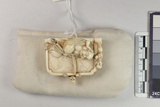 Hearst Museum object 6 of 14 titled Netsuke, accession number 9-18870, described as Netsuke; reclining figure of fat man facing to his right, supporting head on right hand; lying in front of wall; boy at left peering at man; boy at back of wall standing on barrel pulling at kimono cord; boy at right tugging at kimono hem; man holding fan in left hand; both sides of wall visible; H 3.6 cm X L 4.8 cm