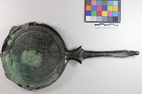 Hearst Museum object 7 of 13 titled Stemmed mirror, accession number 8-5859, described as Etruscan bronze mirror; cast-handle type; encrustation on both surfaces of disk and blue discoloration on obverse due to bronze disease; dark green patina; handle ends in stylized ram’s head; rim of obverse beaded; engraved scene of four figures, two women flanking Minerva (Greek=Athena) and nude man, standing against architecture; guilloche border; mirror appears to have been conserved with a translucent glaze.