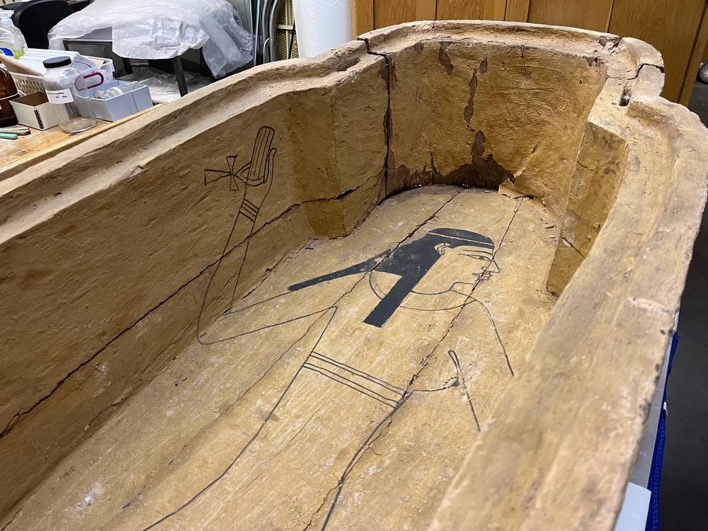 Hearst Museum object 8 of 167 titled Coffin, accession number 6-19912, described as Wooden anthropoid coffin. Inscription running down center of lid. Determined by Kea Johnston to probably be the outer coffin of Iwefaa (6-19928, 12-11039). According to Kea Johnston, "This coffin belonged to a man named Iwefaa, whose name is inserted in the middle of the spells written in the two columns on the front. This coffin is likely the outer coffin for PAHMA 6-19928 based on the similarity of the owner's name, unusual costume, and the paleography of the inscription. Both coffins were purchased on the antiquities market by George Reisner for Phoebe A Hearst. However, stylistically, they are very similar to a set of coffins known to have come from the site of Akhmim, which was excavated in the 1880s by Gaston Maspero, and which has heavily looted for the antiquities market during this decade." "The pectoral vignette between the text and the collar shows the deceased standing before squatting baboon and is the source of our logo. The hieroglyph for 'sky' is above their heads, and the side borders are formed by a pyramid and the symbol for the West.