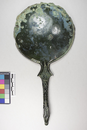 Hearst Museum object 9 of 13 titled Stemmed mirror, accession number 8-5859, described as Etruscan bronze mirror; cast-handle type; encrustation on both surfaces of disk and blue discoloration on obverse due to bronze disease; dark green patina; handle ends in stylized ram’s head; rim of obverse beaded; engraved scene of four figures, two women flanking Minerva (Greek=Athena) and nude man, standing against architecture; guilloche border; mirror appears to have been conserved with a translucent glaze.