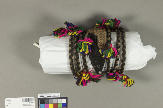 Hearst Museum object titled Sling, accession number 16-20593, described as woven sling of llama of alpaca wool (brown and black) yarn, decorated with small, multicolored (yellow, brown, blue, red, green, pink) pom-poms along length of cords and at either side of woven cup.  cup woven directly into cords using white warp threads, cords end with a pom-pom at one end and a small loop at the other.