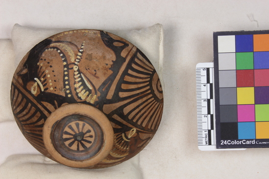 Hearst Museum object 10 of 14 titled Pyxis lid, accession number 8-12a, described as Apulian red figure pyxis lid; several small chips in rim and one in knob; slightly reddened clay; knob glazed with upper edge and central medallion, decorated with rosette, reserved; A-B, head of woman to left, wearing sakkos, diadem, earring, and neckalce; details in black and yellow; between heads, palmettes; strokes on rim. Once in Brussels in the Somzée Collection.