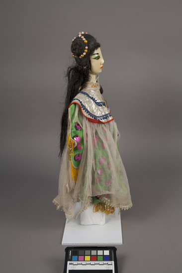 Hearst Museum object titled Hand puppet, accession number 9-22235, described as Puppet, hand.  Foreign Princess A.  Head: carved wood, light orange with pink, green and black features, long, black hair with beads in it  B. Body: red and pink flowered print cloth,  Boots:  wood, black and white.  C. Dress: green cloth with purple sequins, sheer white overlay  48 x 31 cm (18 7/8 x 12 3/16 in)