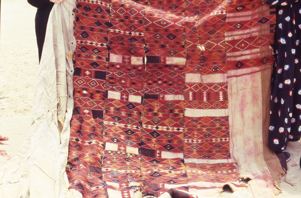 Hearst Museum object titled Color slide, accession number 25-98808, described as 35mm color slide transparency. Multiple people holding up a large textile, pictured below the shoulders. Textile has striped and diamond patterns; colored red, orange, yellow, brown, black, and white.