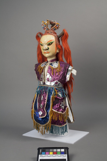 Hearst Museum object 5 of 6 titled Hand puppet, accession number 9-22416, described as Puppet, hand. King.   A.  Head:  carved wood, off-white with orange, green, brown and black features, red-orange hair with 2 high ponytails, crown:  wire wrapped in fabric with pink, blue white and gold beads  B.  Body: white cloth.  Legs:  green-brown cloth.  Hands: wood, white, orange nails.  Boots:  wood, black and white.  C. Robe:  cloth,  metallic purple, silver applique, yellow and blue fringe.  51 x 24 cm (20 1/16 x 9 7/16 in)