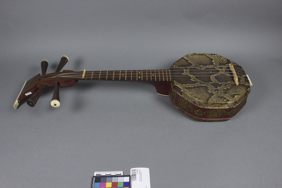 Hearst Museum object titled Long-necked lute, accession number 9-16330, described as Banjo. Metal soundbox with top and bottom covered with snake skin. Neck and pegs of wood, ivory trim. Neck and soundbox painted red with floral trim. Chinese writing on back of neck. Bridge and strings appear to be recent additions. Length: 60.5 cm; soundbox diameter: 19 cm.