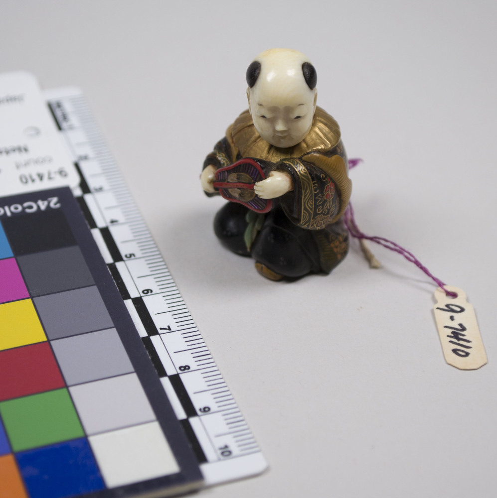 Hearst Museum object titled Netsuke, accession number 9-7410, described as Netsuke: boy with large head, seated. Wears gold collar. Carries fan.