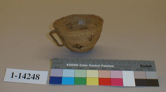 Hearst Museum object titled Teacup, accession number 1-14248, described as Small basket of tea cup model; twined. Warp and weft are twisted Tule skin (Schoenoplectus acutus). White pattern is Reed (Phragmites vulgaris).  Black dye is Tule root. Braided handle of Tule. Small irregular figures in black.