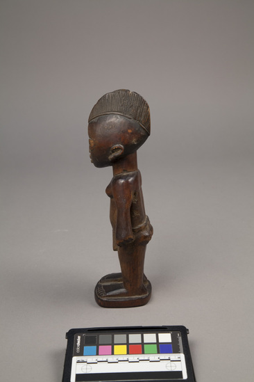 Hearst Museum object 4 of 6 titled Ere ibeji figure, accession number 5-15807, described as Figure, twin (Ibeji), wood, handcarved; female, with elongated headdress, wearing grass skirt; hands on hips; straight legs, with feet mounted on keystone-shaped base; dark brown, polished finish. Bottom stamped "Product of the Gold Coast" by William Bascom in compliance with customs regulation, per collector's notes.