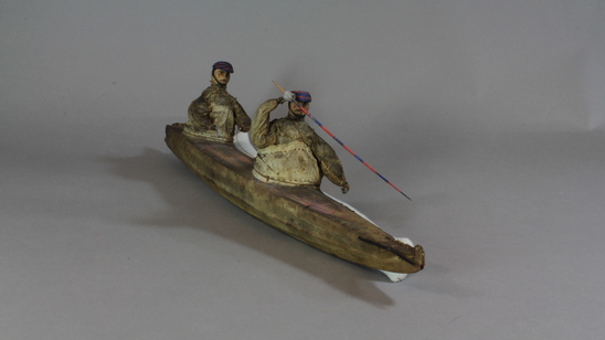 Hearst Museum object 8 of 8 titled Kayak model, accession number 2-4569, described as 2-hatch. Wood frame, rawhide cover. 2 human figures wearing hats painted bright blue and red and wearing sealskin parkas. 15 fixtures including weapons and paddles painted bright red and blue.