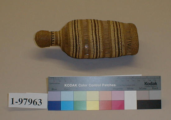 Hearst Museum object 1 of 2 titled Flask, accession number 1-97963, described as Covered with basketry jacket.  Tags "Klamath River tribes unspec.". Per Ralph Shanks:  Twined basketry covered glass liquor bottle with basketry cap.  Rectangular crossed warp starting knot.  The warp material is probably hazel.  The weft material is conifer root.   The weft design overlay is maidenhair fern and beargrass.  Starting after the starting knot, there is 1/4 inch of plain twining followed by 3/4 inch of three strand twining followed by one weft of plain twining.  This is followed by a half inch of parallel warp openwork, followed by a single weft row of plain twining, followed by two weft rows of three strand twining, followed by 1 inch of plain twining, followed by two weft rows of three strand twining, followed by 1/2 inch of parallel openwork, followed by one weft row of plain twining, followed by two weft rows of three strand twining.  This general pattern repeats twice more up to the neck.  After the uppermost openwork there is a single weft row of plain twining followed by a 1/4 inch of three strand twining, followed by a mixture of three strand and plain twining up to the rim.  The cap is stuck on the basket.  The cap has conifer root wefts with no overlay.  The whole cap is done in three strand twining except for a 1/4 inch of plain twining 1/4 inch from the crossed warp start.  The design on the main bottle is horizontal lines of maidenhair fern and beargrass.  The rim is trimmed.  Both of the baskets have a rightward work direction, with an up to the right slant of weft twist. The workface is on the exterior.  The basket is from Northwestern California.