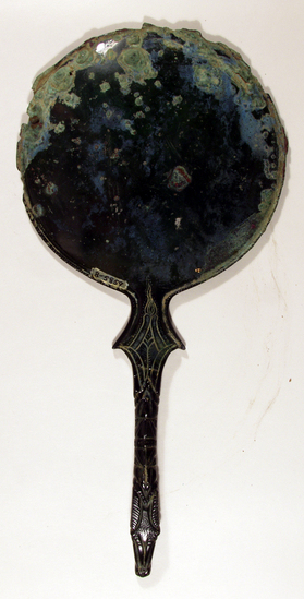 Hearst Museum object 5 of 13 titled Stemmed mirror, accession number 8-5859, described as Etruscan bronze mirror; cast-handle type; encrustation on both surfaces of disk and blue discoloration on obverse due to bronze disease; dark green patina; handle ends in stylized ram’s head; rim of obverse beaded; engraved scene of four figures, two women flanking Minerva (Greek=Athena) and nude man, standing against architecture; guilloche border; mirror appears to have been conserved with a translucent glaze.