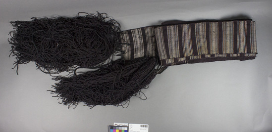 Hearst Museum object titled Sash, accession number 7-5123, described as Woman's wool sash (pojas) made from homespun; brownish-purple; white and blue weft designs near ends; very long fringe; 5" wide, 142" long. Used by women for everyday wear as sash under prepaska sash, 7-5124. Worn alone by older women. Fringe at end known as amajlija, "amulet". Made by women for own use and still worn by some women at time of collection. 7-5119-5128 comprise a complete costume.