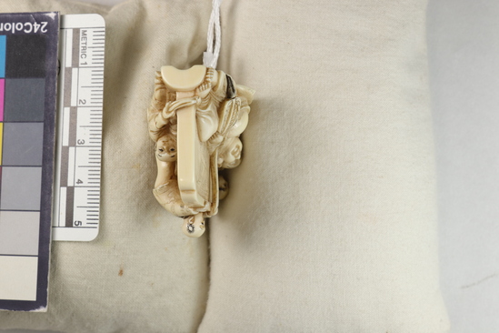 Hearst Museum object 11 of 14 titled Netsuke, accession number 9-18870, described as Netsuke; reclining figure of fat man facing to his right, supporting head on right hand; lying in front of wall; boy at left peering at man; boy at back of wall standing on barrel pulling at kimono cord; boy at right tugging at kimono hem; man holding fan in left hand; both sides of wall visible; H 3.6 cm X L 4.8 cm