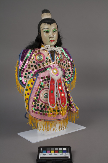 Hearst Museum object 5 of 10 titled Hand puppet, accession number 9-22260, described as Puppet, hand.  Young Man.  A.  Head:  wood, carved, white and pink with green, pink and black features, black hair in bun.  B.  Body:  cloth, brown.  Legs:  cloth, green and pink.  Hands:  wood, white.  Boots:  wood, black and white.  C. Robe:  cloth, pink with green, pink and gold sequins.  yellow fringe.  50 x 36 cm (19 11/16 x 14 3/16 in).