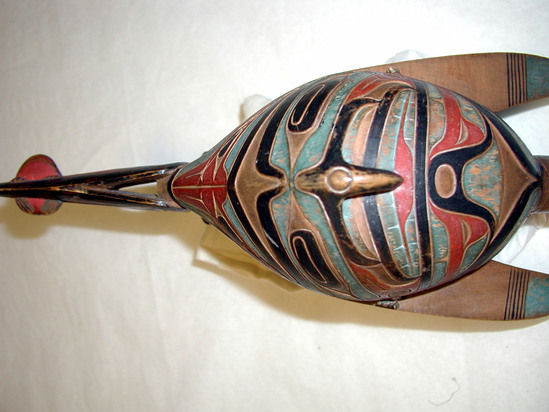 Hearst Museum object 6 of 9 titled Rattle, accession number 2-19094, described as Totemic wooden rattle in raven form, human being on back with frog on his stomach and hawk head behind his legs, raven "ears" connect with head of human figure, sparrow hawk carving on underside; alder; painted red, blue, and black. Frog sitting on human figure is sticking its tongue into mouth of human. Raven head with frog in mouth refers to a myth where raven took frog in its mouth and descended to the depths of the sea, frog acting as lungs for raven. Raven, frog and wood identified by Charles Brown.