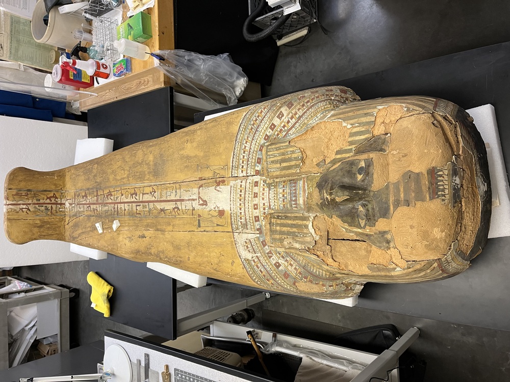 Hearst Museum object 148 of 167 titled Coffin, accession number 6-19912, described as Wooden anthropoid coffin. Inscription running down center of lid. Determined by Kea Johnston to probably be the outer coffin of Iwefaa (6-19928, 12-11039). According to Kea Johnston, "This coffin belonged to a man named Iwefaa, whose name is inserted in the middle of the spells written in the two columns on the front. This coffin is likely the outer coffin for PAHMA 6-19928 based on the similarity of the owner's name, unusual costume, and the paleography of the inscription. Both coffins were purchased on the antiquities market by George Reisner for Phoebe A Hearst. However, stylistically, they are very similar to a set of coffins known to have come from the site of Akhmim, which was excavated in the 1880s by Gaston Maspero, and which has heavily looted for the antiquities market during this decade." "The pectoral vignette between the text and the collar shows the deceased standing before squatting baboon and is the source of our logo. The hieroglyph for 'sky' is above their heads, and the side borders are formed by a pyramid and the symbol for the West.