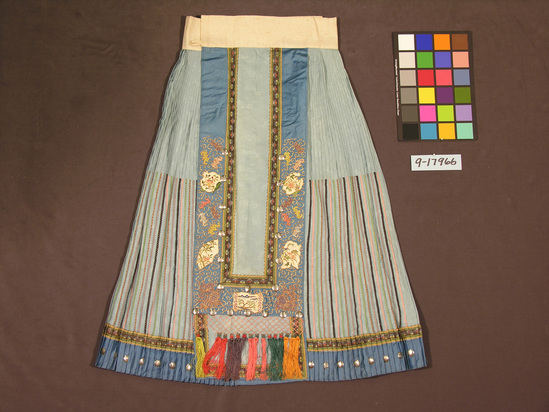 Hearst Museum object titled Skirt, accession number 9-17966, described as Skirt:  women's pale blue silk gauze patterned w/ bats & lozenges; lower portion of pleated panels decorated w/ narrow bands of multicolor woven tape; wide front & back panels bordered w/ wide blue satin bands couched w/ gold-wrapped tread & appliqued white & purple satin cutouts embroidered in floral motifs; multicolor macrame work & tassels line bottom of wide panels; silver metal disks sewn around blue satin hem & edge wide panels; waist band w/ loop & toggle closures in plain white cotton.  Note: white satin appliques probably predate garment.   Women's skirt; c. 1900, applique c. 1850, fide Mary Hays.  92.5 cm L.