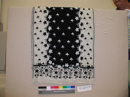 Hearst Museum object titled Shawl, accession number 9-21513, described as Shawl; 2 x 2 twill weave; pieced from 5 pieces of cloth- 3 lengthwise &1 at each end. Machine stitched seams covered with embroidery; white with large black inset center; floral black and white chainstitch embroidery- black on white ground and white on black ground;  embroidered patterns include grapes and leaves at each end, leaf motifs throughout, and stylized floral border along each selvage; 1 cloth selvage handseamed, 1 woven selvage; warps are grouped and tied in bundles to form decorative fringe, except for 1 cm woven strip at each selvage.