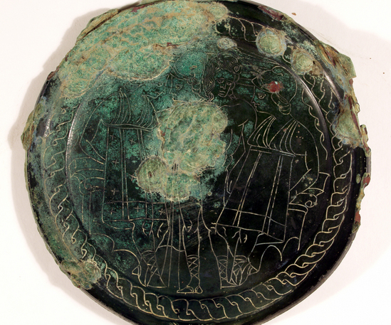 Hearst Museum object 10 of 13 titled Stemmed mirror, accession number 8-5859, described as Etruscan bronze mirror; cast-handle type; encrustation on both surfaces of disk and blue discoloration on obverse due to bronze disease; dark green patina; handle ends in stylized ram’s head; rim of obverse beaded; engraved scene of four figures, two women flanking Minerva (Greek=Athena) and nude man, standing against architecture; guilloche border; mirror appears to have been conserved with a translucent glaze.