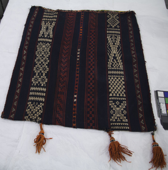 Hearst Museum object titled Bag, accession number 9-23737, described as Storage bag, khurj. Dark blue predominating with red and white. Has wide bands of saha weave in blue/white and blue/red. 'Weirjan near outer edges. Saha patterns are in solids, typical or northern style. Patch near top edge, using binding cloth. S-ply. Blue may be Indigo and red madder. Darkness of color suggests at least partial use of natural dye. Warp-faced weave with warp substitution. 12 EPI thread count per inch. Edges sewn together with rough chevron stitch in maroon. Tassels: 4 worn, tattered orange ones. Center seam handsomely sewn in faggotting stitches in blue, red, orange, and white blocks. Top edge crudely bound in print cloth on one side, totally worn out red over sewn in blue wool on the other side.