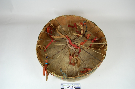 Hearst Museum object 3 of 5 titled Drum and drumstick, accession number 2-66139a,b, described as a) Drum.  Single head, rawhide over wood hoop frame.  Strips of dyed red and blue cotton cloth laced around eges, short strips of of same cloth are tied to framework of the underside; blue glass crow beads on three strips.  b) Carved wood drumstick.