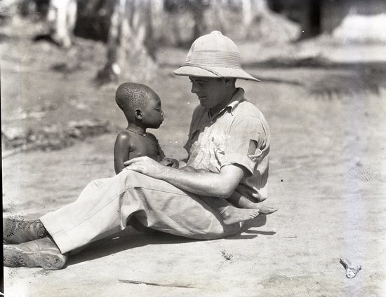 Hearst Museum object titled Black and white negative, accession number 15-32435, described as 4" x 5" Negative. Harold Austin sits down and converses with a small child in Monvu.