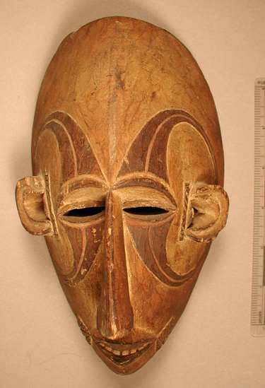 Hearst Museum object titled Mask, accession number 5-16143, described as Ibo Mask. carved wood, stained designs, representing beautiful woman. natural wood skin color, large nose, narrow slit eyes, carved ears. protuding lips with bared, incised teeth. 6 on top, 5 on bottom. fan shaped design extending from each corner of mouth. 2 circular designs on each side of face, circle extending from eyes, to mid-forehead, around ears, to mid cheek, back around to eyes.  Ibo Mask (Udi, 2, 700, 1945). Fine Beautiful women, old. 8"1/4"L, 4-3/4"W