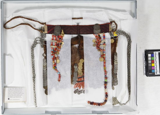 Hearst Museum object titled Face veil, accession number 5-16986, described as Face covering with sheer, brown crepe veil - decorated with 16 bands of silver coins on each side and 5 gold colored bangles plus a pendant of coral beads and silver coins at center. Brow band (length: 31.0 cm) is cross-stitched with silk thread (black diagonal, open diamonds on a red ground) is attached to the veil at center and via a double chain at each temple. Band is decorated on each side with 2 large silver coins, gold colored amulets (1 missing) and 6 "tassels" of beads of varying lengths (5.5 cm to 43.0 cm). A braided band attached to the brow band ties around the head, 3 silver colored chains are attached at both temples of veil, one terminating with silver, diamond shaped pendant, decorated with repoussé stars and half moons. Veil fabric smocked at top and finished with bands of cording at top. Bead tassels include coral, turquoise, glass, plastic, shell, wood, pearl and mother of pearl beads. From North Sinai, Egypt.