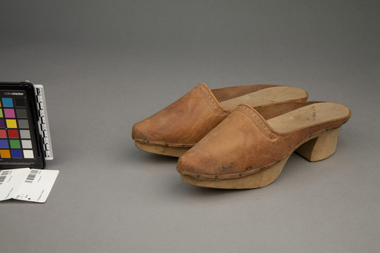 Hearst Museum object titled Slippers, accession number 10-76a,b, described as Slippers with wooden soles and heels and leather tops; no back; connecting strap broken; tooled design on tops; 11 inches long.