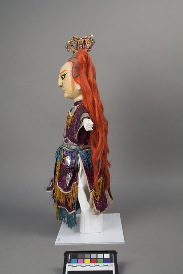 Hearst Museum object 3 of 6 titled Hand puppet, accession number 9-22416, described as Puppet, hand. King.   A.  Head:  carved wood, off-white with orange, green, brown and black features, red-orange hair with 2 high ponytails, crown:  wire wrapped in fabric with pink, blue white and gold beads  B.  Body: white cloth.  Legs:  green-brown cloth.  Hands: wood, white, orange nails.  Boots:  wood, black and white.  C. Robe:  cloth,  metallic purple, silver applique, yellow and blue fringe.  51 x 24 cm (20 1/16 x 9 7/16 in)