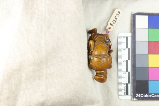 Hearst Museum object titled Netsuke, accession number 9-12537, described as Carving / Cabinet piece (?) or possibly Netsuke: Carved ivory figure of walking man, left leg raised in step. His face is turned to the left, and is smiling. Slung on his right side is a bag w/ three characters written upon it, a small inlaid piece of coral, and an inlaid mother of pearl 'double gourd'. The entire right hand is broken off, as are the tips of the fingers of the left hand and right toes. Cloud designs incised on the back of the garment. H. 4.5 cm.