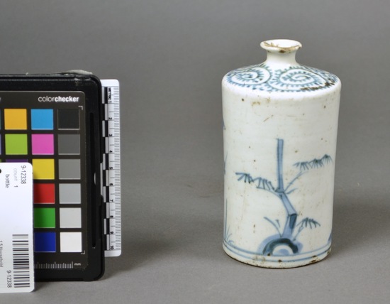 Hearst Museum object titled Bottle, accession number 9-12338, described as Bottle, porcelain. Cylindrical shape with almost horizontal top to small narrow neck. Small flared mouth. Underglaze blue decoration in varied tone strengths, of bamboo with leaves and stylized fruit tree blossoming, separated by grass on each side. Two bands below near base. Shoulder has octopus pattern on shoulder-top. Rim is chipped and worn, many spots on body. Recessed foot has crack. Height 13.5 cm, Base diameter 6.5cm. From Japan, Saga Prefecture (Hizen), Arita, "Imari" ware.