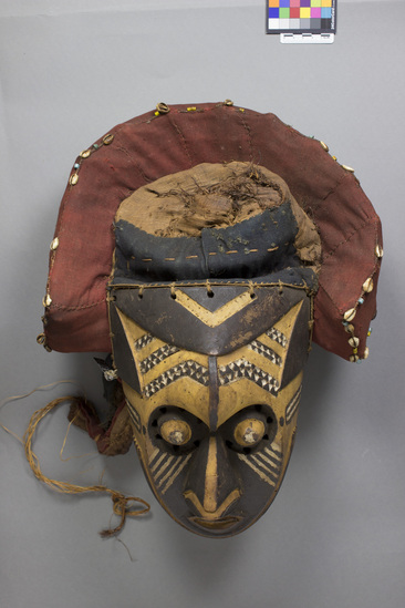 Hearst Museum object titled Wood/fabric mask, accession number 5-12967, described as Mask; anthropomorphic mask with prominent triangular nose, bulging eyes perforated inside outer perimeter; prominent forehead; yellow ground with black and white pigmentation; diagonal slashes on cheeks; separately carved ears attached behind on fiber-lined red-stained cloth decoration which is above mask; cowries and trade beads embellish the cloth; height of face only ca. 26 cm