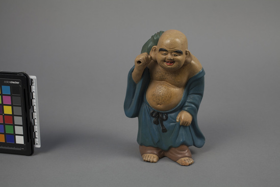 Hearst Museum object titled Figurine, accession number 9-4873, described as Pottery figure of standing Hotei, one of the Seven Gods of Fortune; drabbish and blue, 7  3/4” high.