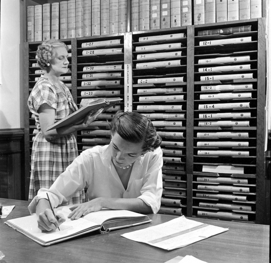 Hearst Museum object titled Black-and-white print, accession number 13-3076, described as R. Kay Eckman & M. Laferriere in the office of the UC Museum of Anthropology, entering cataloging information into ledgers.