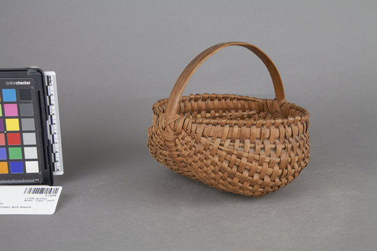 Hearst Museum object titled Basket, accession number 2-72559, described as Woven, willow basket with wooden handle, base support and rim reinforcement.  Weft pieces are of tapered wooden pieces.  Light brown of variegated shades.  Base support forms a slightly recessed center, thus the 2 sides of the basket form lobes giving it a distinct shape.