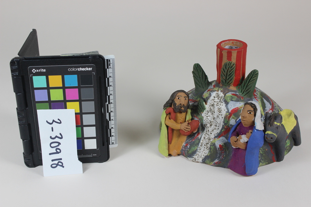 Hearst Museum object titled Candlestick, accession number 3-30918, described as CANDLE STICK:  Ceramic candlestick formed from a half globe, to which a hollow tube for mounting a candle is attached at top.  Attached at the base, on one side, are a woman in a purple gown, blue cloak and white head cover holding a swaddled infant; a bearded man wearing a yellow ochre robe, yellow cloak and holding a bottle and shepherds crook;  and a grey donkey with and a yellow saddle.  The globular base is painted green, grey, yellow and red.  A white stream flowing down from the candle stick to the edge of the base, and highlighted with silver glitter, between the two standing figures.  Three green trees flank the candleholder at top.  Holder is painted with red stripes.  Appears to represent the flight into Egypt by the Christian holy family.  “Mexico” stamped on underside.