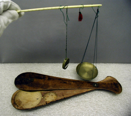 Hearst Museum object 1 of 3 titled Scale, accession number 9-21344a-c, described as Pair of scales in wood case. Brass weight (c) hanging by green silk cord from calibrated ivory or walrus-tooth beam. Bowl (a) of the same metal for pinched-off silver. Beam (also a) to be held by one of three green silk threads, according to weight of silver. Center thread with red tassel. Closure of wood case (b) with copper(?) latch; rivet also copper(?). Length of beam: 25 cm; width of bowl: 6 cm; length of case: 29 cm; greatest width: 7 cm.