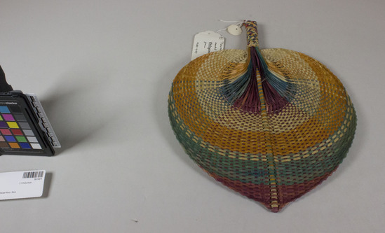 Hearst Museum object titled Fan, accession number 18-1471, described as Fan (decorated). Magenta, red, gold, natural fiber color. Checkerboard effect on cylindrical handle, loop on handle. Leaf shape with central rod for support. Plain weave for most of fan, except “heart” which is not woven, but single strands giving peacock effect. Length 36 cm. Width 27 cm.