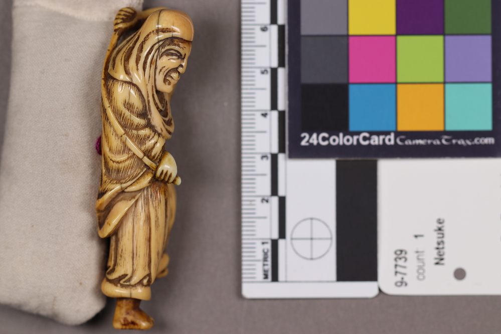 Hearst Museum object 3 of 9 titled Netsuke, accession number 9-7739, described as Netsuke: tall thin man with long face. Left foot raised, holds long curved rod behind his back.
