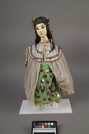 Hearst Museum object 2 of 6 titled Hand puppet, accession number 9-22235, described as Puppet, hand.  Foreign Princess A.  Head: carved wood, light orange with pink, green and black features, long, black hair with beads in it  B. Body: red and pink flowered print cloth,  Boots:  wood, black and white.  C. Dress: green cloth with purple sequins, sheer white overlay  48 x 31 cm (18 7/8 x 12 3/16 in)