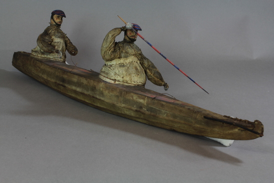 Hearst Museum object 4 of 8 titled Kayak model, accession number 2-4569, described as 2-hatch. Wood frame, rawhide cover. 2 human figures wearing hats painted bright blue and red and wearing sealskin parkas. 15 fixtures including weapons and paddles painted bright red and blue.