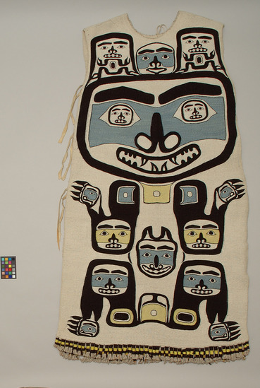 Hearst Museum object 3 of 3 titled Tunic, accession number 2-59550, described as Abstracted bear designs; black, yellow and blue on white; dyed mountain goat wool twined over wool-covered cedar bark warp; front design is standing bear with large face; smaller faces within ears and at joints; on back are (top to bottom) a pair of eyes, 4 spinal vertebrae?, and black/yellow checkerboard panel; tassels at base of garment; one side of tunic is sewn almost closed, other side has 3 pair buckskin ties; drawstring at neck. Designs said to be grizzly bear and 2 cubs.